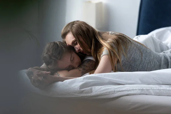 Enfoque selectivo de dos lesbianas cogidas de la mano y abrazando mientras yacen en la cama por la mañana - foto de stock
