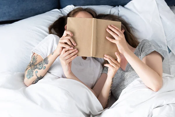 Dos lesbianas acostadas debajo de una manta y sosteniendo el libro en la cama - foto de stock