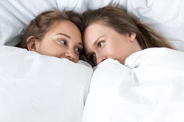 Due lesbiche sdraiate sotto una coperta bianca e che si guardano in camera da letto — Foto stock