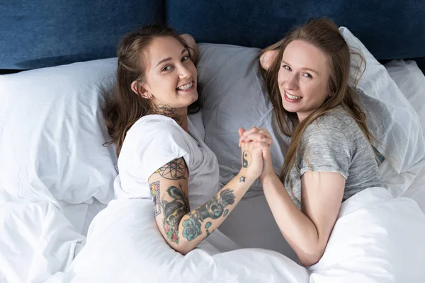 Two laughing lesbians holding hands while lying on bed in morning — Stock Photo