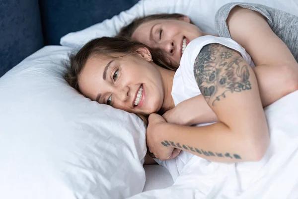Dos lesbianas sonrientes abrazándose mientras yacen en la cama - foto de stock