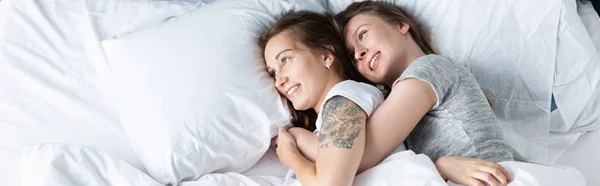 Tiro panorâmico de duas lésbicas sorridentes abraçando enquanto deitado na cama — Fotografia de Stock
