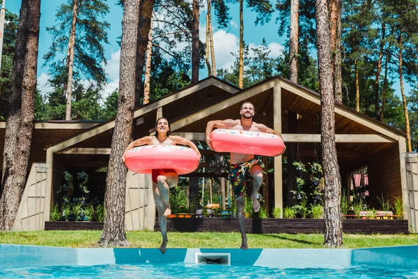 Alegre hombre y mujer con anillos inflables saltando en la piscina - foto de stock