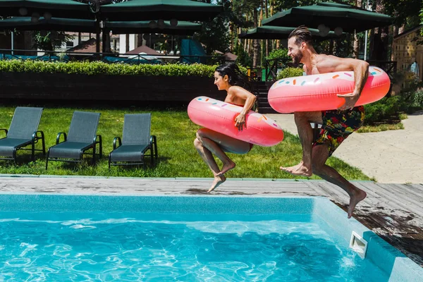 Mujer alegre y hombre con anillos inflables rosados saltando en la piscina - foto de stock