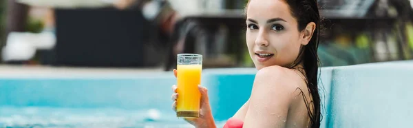 Panoramic shot of happy brunette girl holding glass of orange juice in swimming pool — Stock Photo