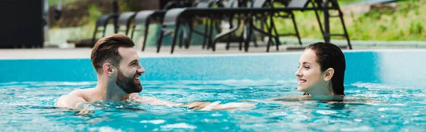 Plano panorámico de hombre barbudo guapo sonriendo mientras mira a la atractiva mujer en la piscina - foto de stock