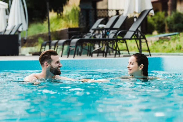 Foyer sélectif de bel homme barbu souriant tout en regardant la femme attrayante dans la piscine — Photo de stock