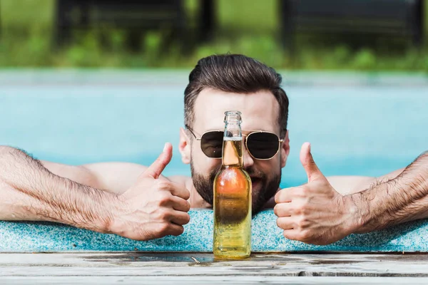 Hombre barbudo en gafas de sol mostrando los pulgares hacia arriba cerca de la botella con cerveza - foto de stock