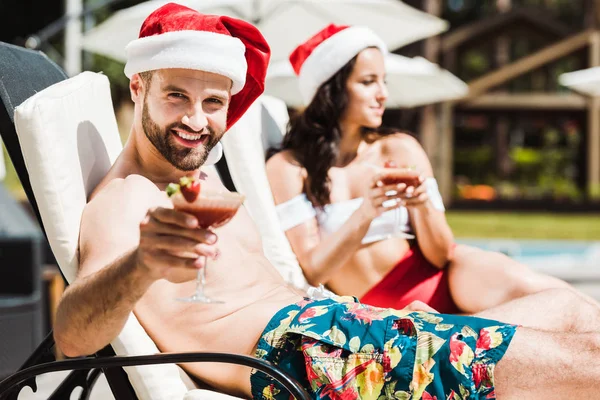 Избирательный фокус счастливого бородатого мужчины в шляпе Санта-Клауса, держащего коктейльный напиток рядом с женщиной — стоковое фото