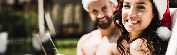 Panoramic shot of happy woman in santa claus hat and man holding sparklers — Stock Photo