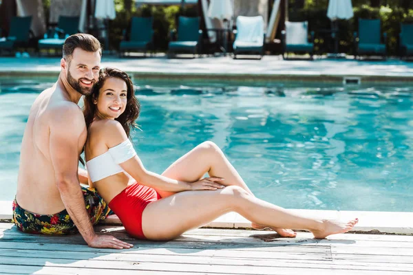 Happy bearded man sitting with attractive woman on wooden decks near swimming pool — Stock Photo