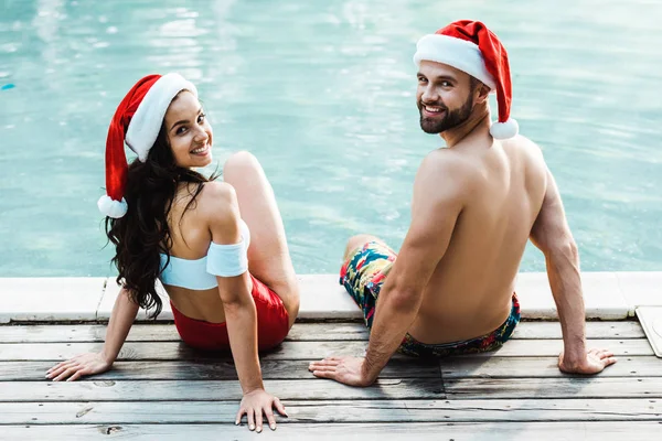 Cheerful man and woman in santa claus hats sitting on wooden decks near swimming pool — Stock Photo