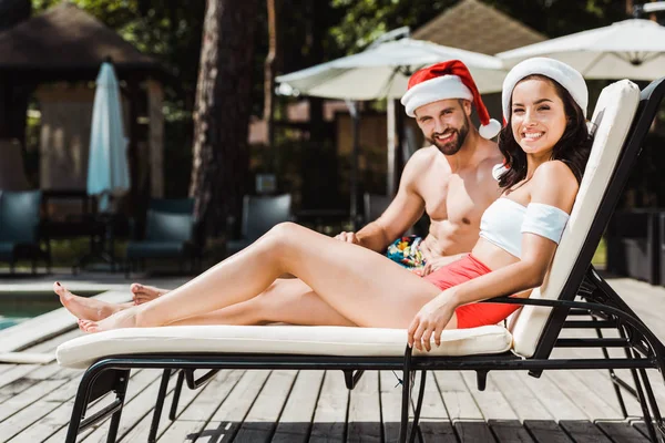 Cheerful man and woman in santa claus hats sitting on wooden deck chairs — Stock Photo