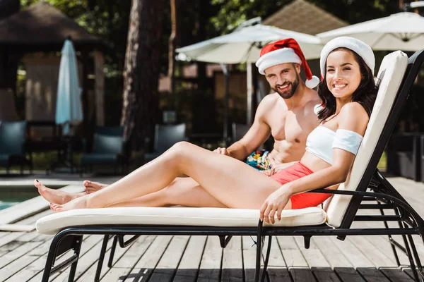 Homme et femme joyeux chapeaux santa claus assis sur des chaises longues en bois — Photo de stock