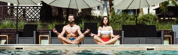 Panoramic shot of woman and handsome man meditating near swimming pool — Stock Photo