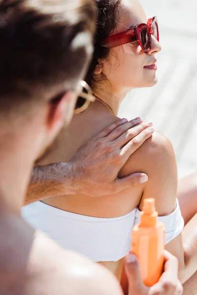 Vista recortada del hombre tocando a la mujer en gafas de sol y sosteniendo la botella con protector solar - foto de stock