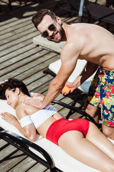 Selective focus of happy man holding bottle with sunblock and touching attractive woman lying on deck chair — Stock Photo