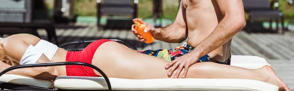 Panoramic shot of man touching young woman lying on deck chair and holding bottle with sunblock — Stock Photo