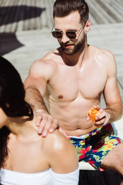 Foco seletivo de homem bonito em óculos de sol tocando jovem e segurando garrafa com protetor solar — Fotografia de Stock