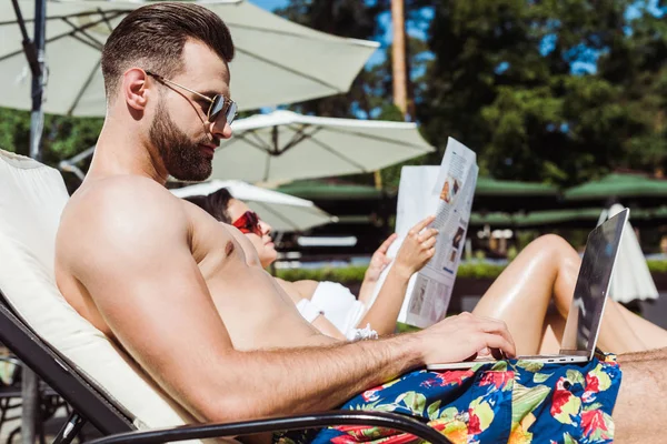 Селективное внимание мужчины в солнцезащитных очках с помощью ноутбука рядом с женщиной с газетой — стоковое фото