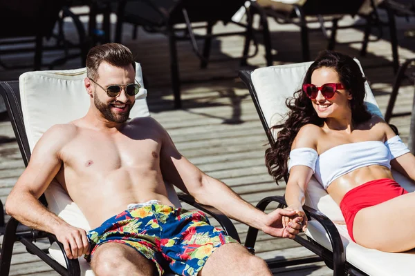 Homem feliz e mulher em óculos de sol de mãos dadas enquanto deitado em cadeiras de convés — Fotografia de Stock