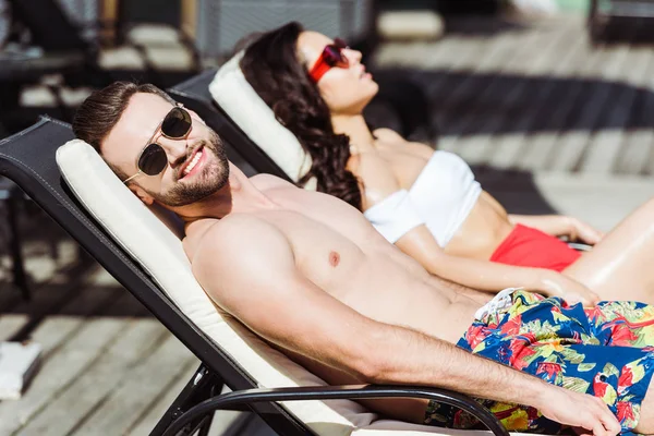 Foco seletivo do homem muscular feliz em óculos de sol deitado na cadeira de praia perto da mulher — Fotografia de Stock