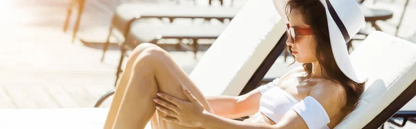 Панорамный снимок привлекательной и стильной женщины в соломенной шляпе и солнцезащитных очках, сидящей на шезлонге — стоковое фото