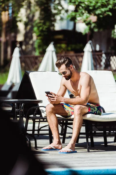 Selective focus of handsome bearded man sitting on deck chair and using smartphone — Stock Photo