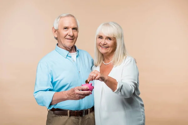 Homme retraité gai tenant tirelire rose tandis que la femme aînée mettre pièce isolée sur beige — Photo de stock