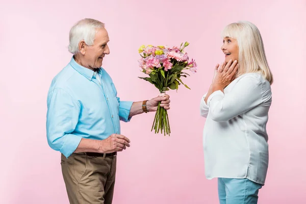 Веселый пожилой мужчина дарит букет с цветами взволнованной жене на розовом — стоковое фото