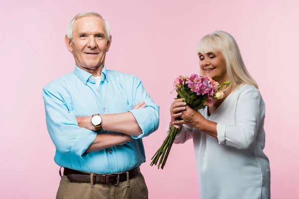 Selektiver Fokus eines glücklichen älteren Mannes mit verschränkten Armen in der Nähe einer glücklichen Frau mit Blumen auf rosa — Stockfoto