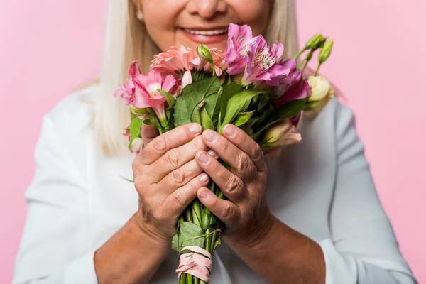 Vista recortada de mujer mayor positiva sosteniendo ramo de flores en flor aislado en rosa - foto de stock