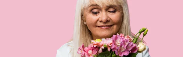 Panoramaaufnahme einer glücklichen Seniorin, die einen Strauß blühender Blumen betrachtet, isoliert auf rosa — Stockfoto