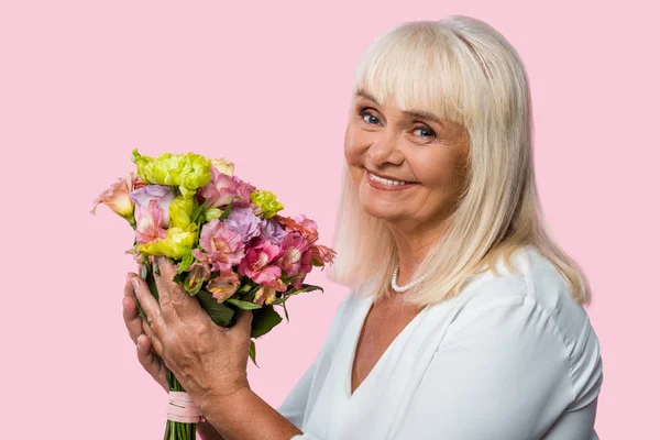 Счастливая пожилая женщина держит цветущие цветы изолированы на розовый — стоковое фото