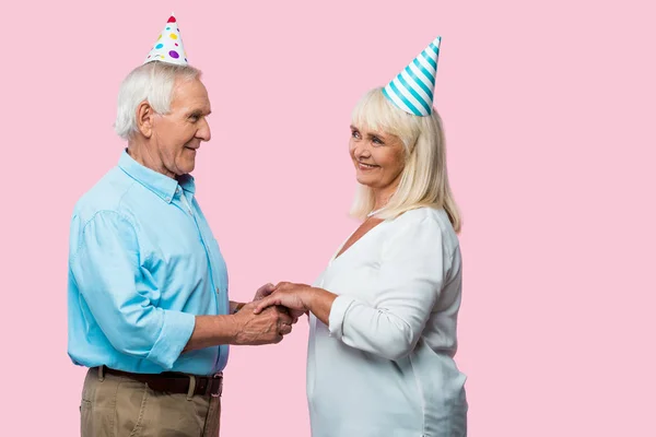 Feliz hombre mayor cogido de la mano con la esposa en la gorra del partido aislado en rosa - foto de stock