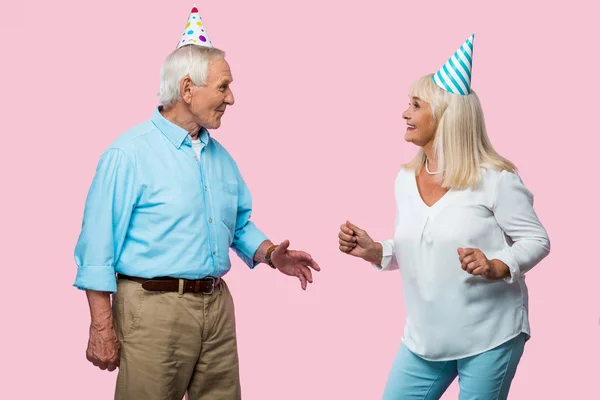 Happy senior woman gesturing and looking at cheerful husband in party cap isolated on pink — Stock Photo