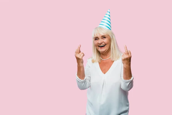 Cheerful senior woman in party cap showing middle finger isolated on pink — Stock Photo