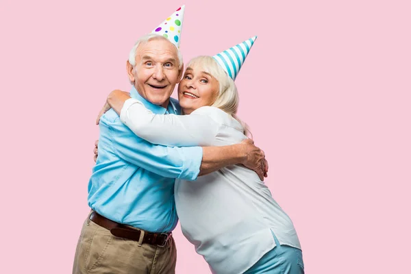 Cheerful retired husband and wife in party caps hugging isolated on pink — Stock Photo