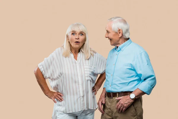 Alegre hombre mayor mirando sorprendido esposa de pie con las manos en las caderas aislado en beige - foto de stock
