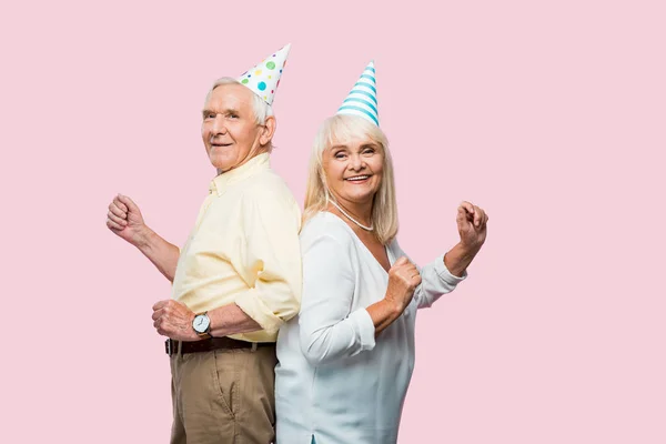 Cheerful senior husband and wife in party caps gesturing isolated on pink — Stock Photo