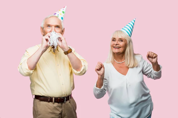 Старший чоловік вкриває обличчя вечірнім вентилятором біля веселої дружини в партійній кепці жестикулює ізольовано на рожевому — стокове фото