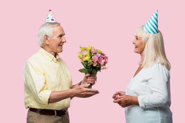 Senior hombre dando ramo de flores a feliz esposa en partido gorra aislado en rosa - foto de stock