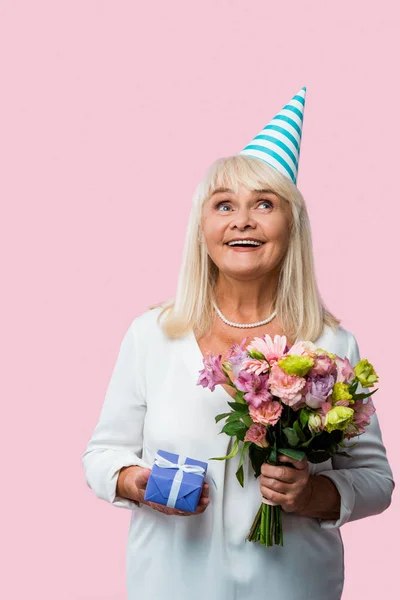 Щаслива старша жінка у вечірній кепці тримає квіти та подарункову коробку ізольовано на рожевому — стокове фото