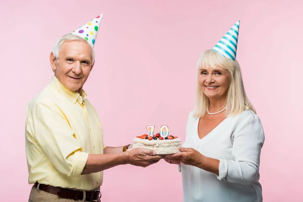 Feliz pareja de ancianos en gorras de fiesta celebración de pastel de cumpleaños aislado en rosa - foto de stock