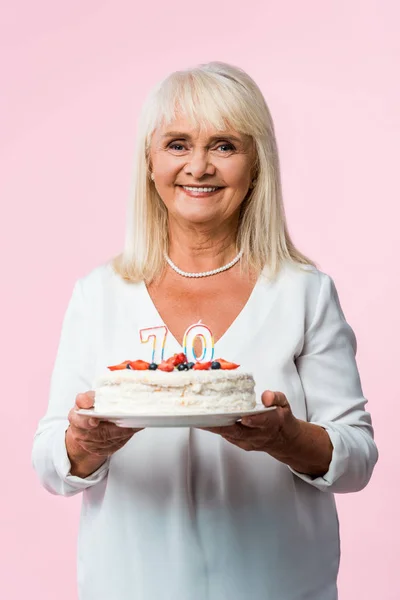 Happy senior woman with grey hair holding tasty birthday cake isolated on pink — Stock Photo