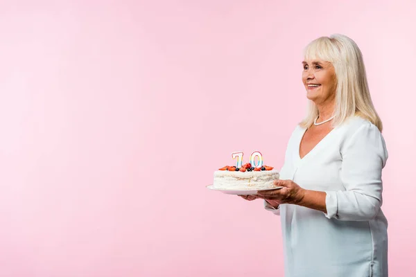 Счастливая женщина в отставке с седыми волосами проведение вкусный торт день рождения изолирован на розовый — стоковое фото
