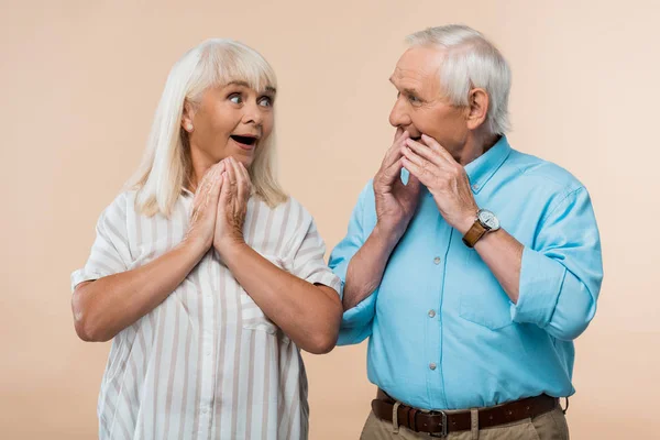 Sorprendida mujer mayor mirando a su marido cubriendo la cara con las manos aisladas en beige - foto de stock
