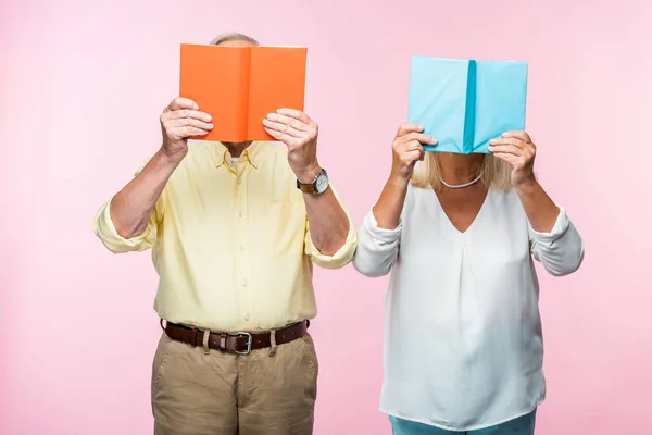 Pareja de pensionistas de pie y cubriendo caras con libros aislados en rosa - foto de stock