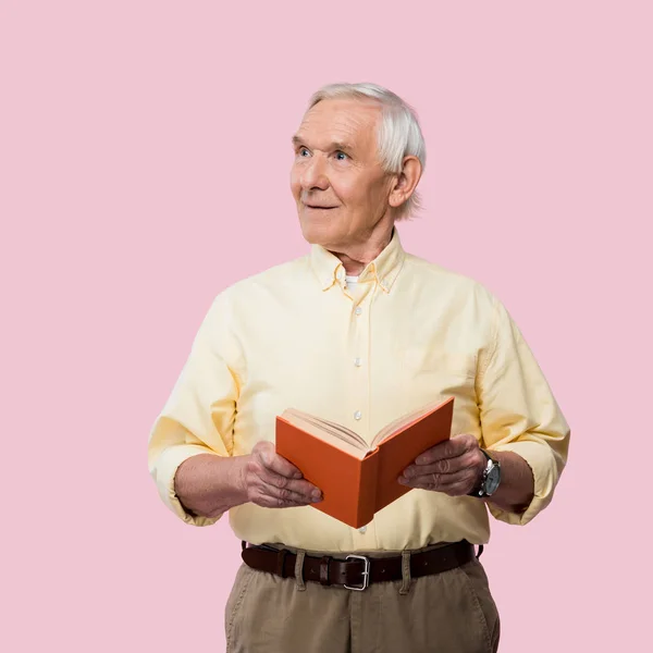 Fröhlicher Rentner hält Buch in der Hand und lächelt isoliert auf rosa — Stockfoto