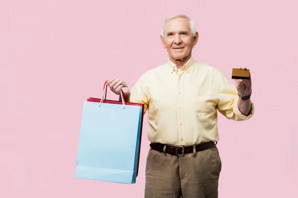 Happy retired man holding credit card and shopping bags isolated on pink — Stock Photo